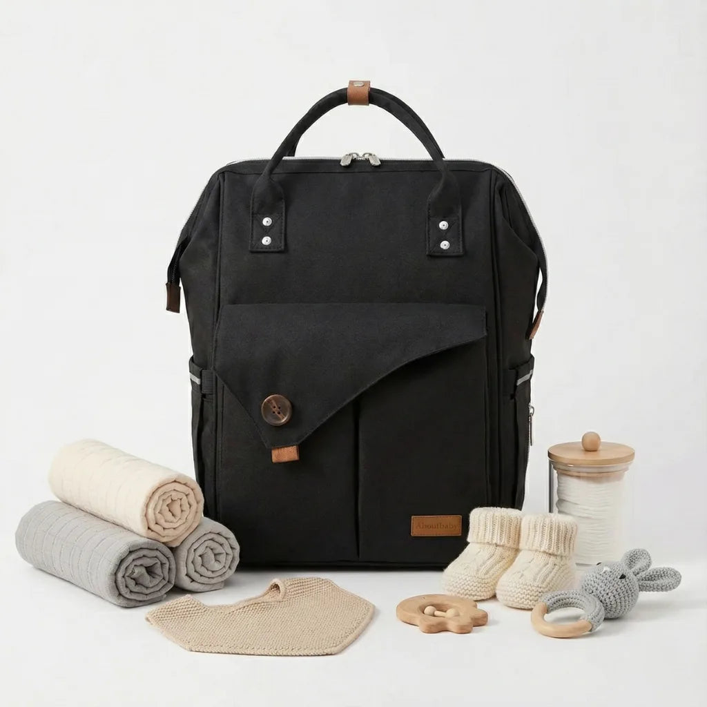 Sac à langer à dos urbain 'Urbannest' noir, grande capacité avec poches multiples, accessoires bébé neutres assortis (langes, chaussons, jouet en bois, doudou lapin) sur fond blanc studio.