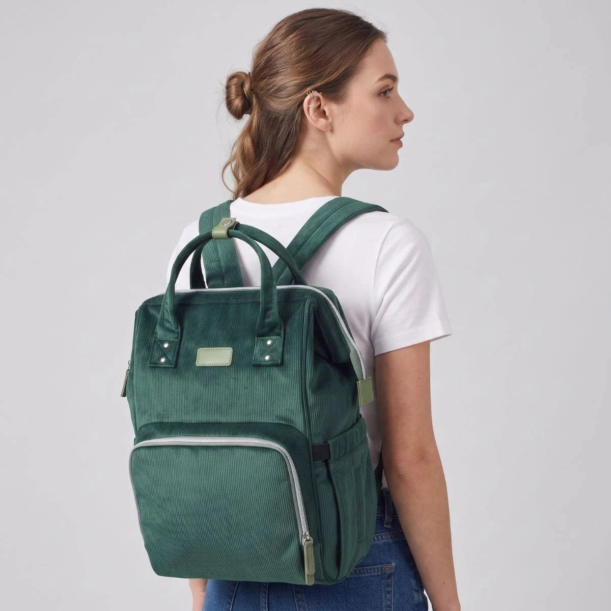 Jeune femme de dos portant le sac à dos à langer Goodnvibes.com en velours côtelé vert émeraude, style minimaliste premium, format 45x30x22cm avec matelas à langer inclus.