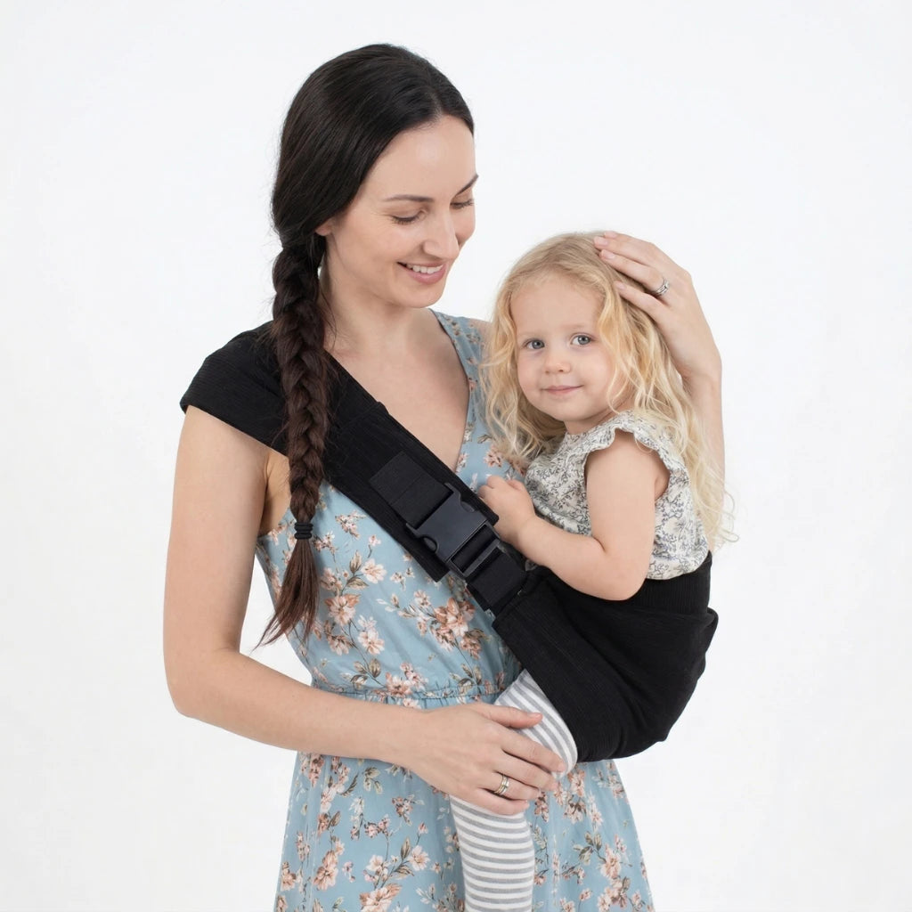 Une maman porte sa petite fille dans une écharpe de portage ergonomique noire, asymétrique avec une grande boucle noire. La maman sourit en caressant tendrement la tête de sa fille bouclée.