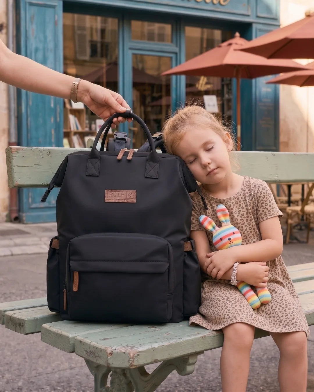 Sac à dos à langer noir Nyla Goodnvibes grande capacité posé sur un banc public avec une petite fille reposant sa tête dessus.