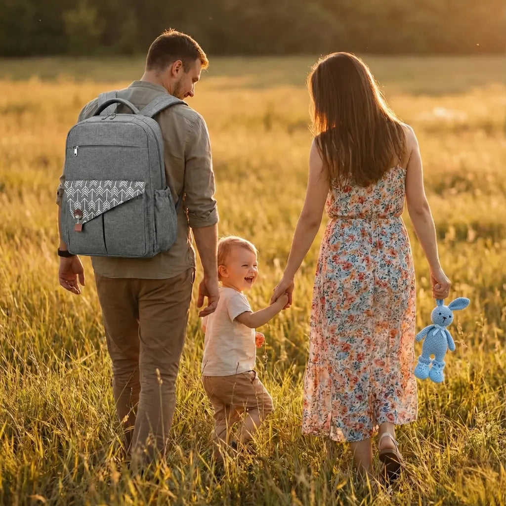 Sac à dos à langer gris texturé avec poches, rabat avant à motifs flèches blanches, accessoires bébé assortis (couches, lange, brosse, jouets en bois) sur fond blanc.