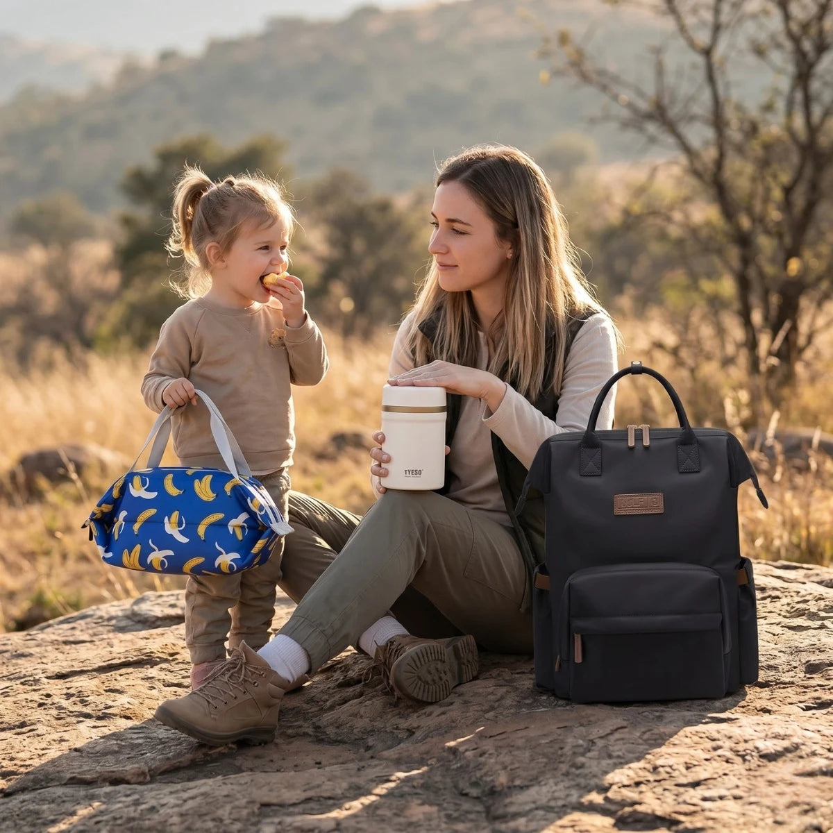 Maman et son enfant en extérieur profitant d'un repas avec le sac à dos à langer Nyla noir déperlant, le lunch bag isotherme FreshBox motif banane et la bento box FreshMeal.