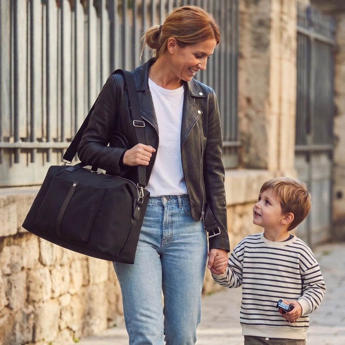 Maman marchant en ville avec son enfant portant le sac à langer MoodPack noir à l'épaule. Sac de maternité pratique et élégant par Goodnvibes